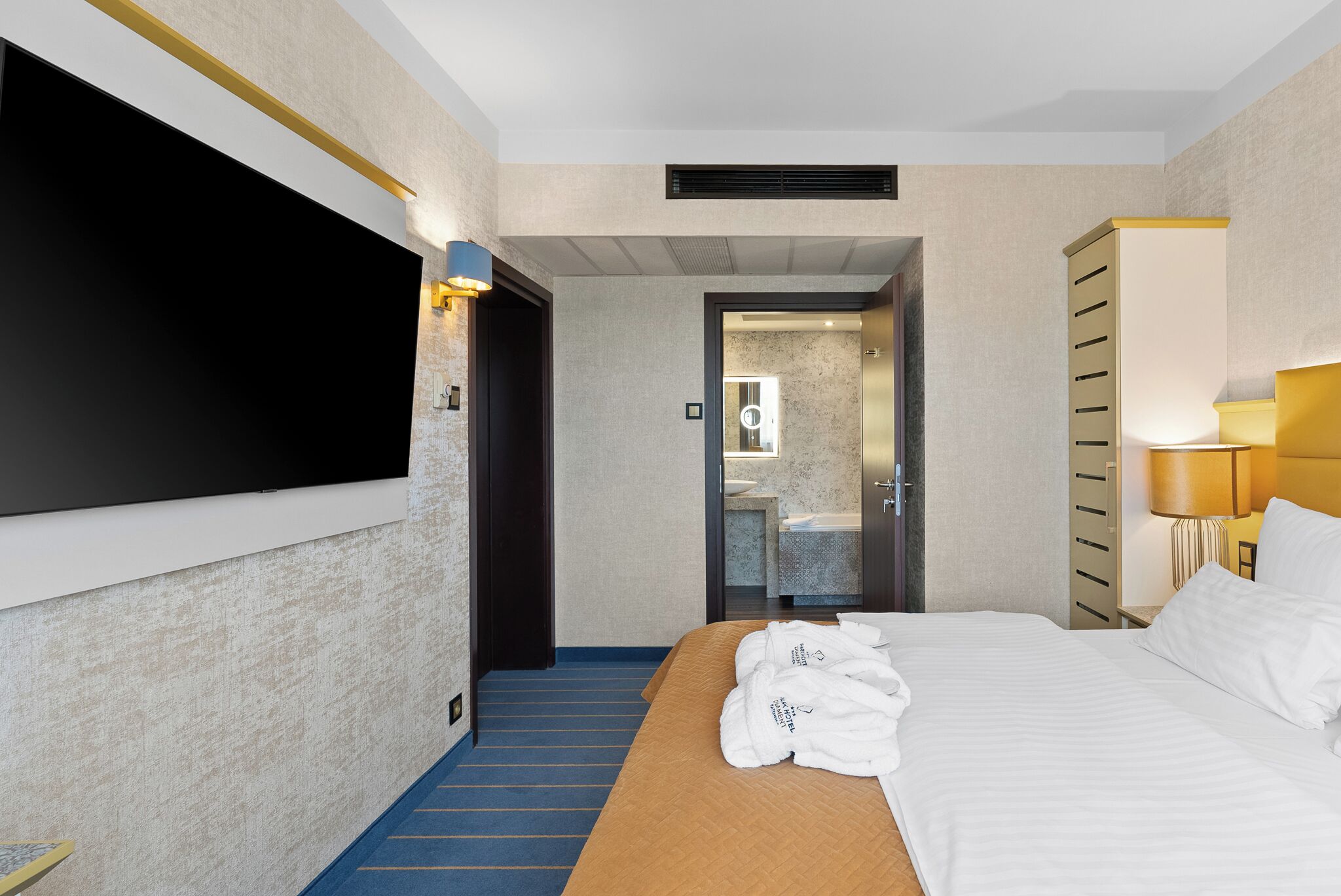 Una habitación de hotel con cama, televisor montado en la pared, baño visible a través de una puerta abierta y una mesita de noche con lámpara.