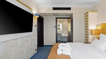 A hotel room with a bed, a TV mounted on the wall, a bathroom visible through an open door, and a bedside table with a lamp.