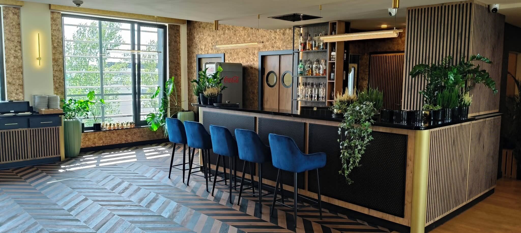 Un espace bar avec un sol décoré d’un motif en chevrons, des tabourets de bar bleus et un comptoir agrémenté de plantes et de bouteilles.