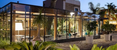 A modern glass-enclosed outdoor dining area with tables and chairs, surrounded by lush greenery and a stone-paved floor.