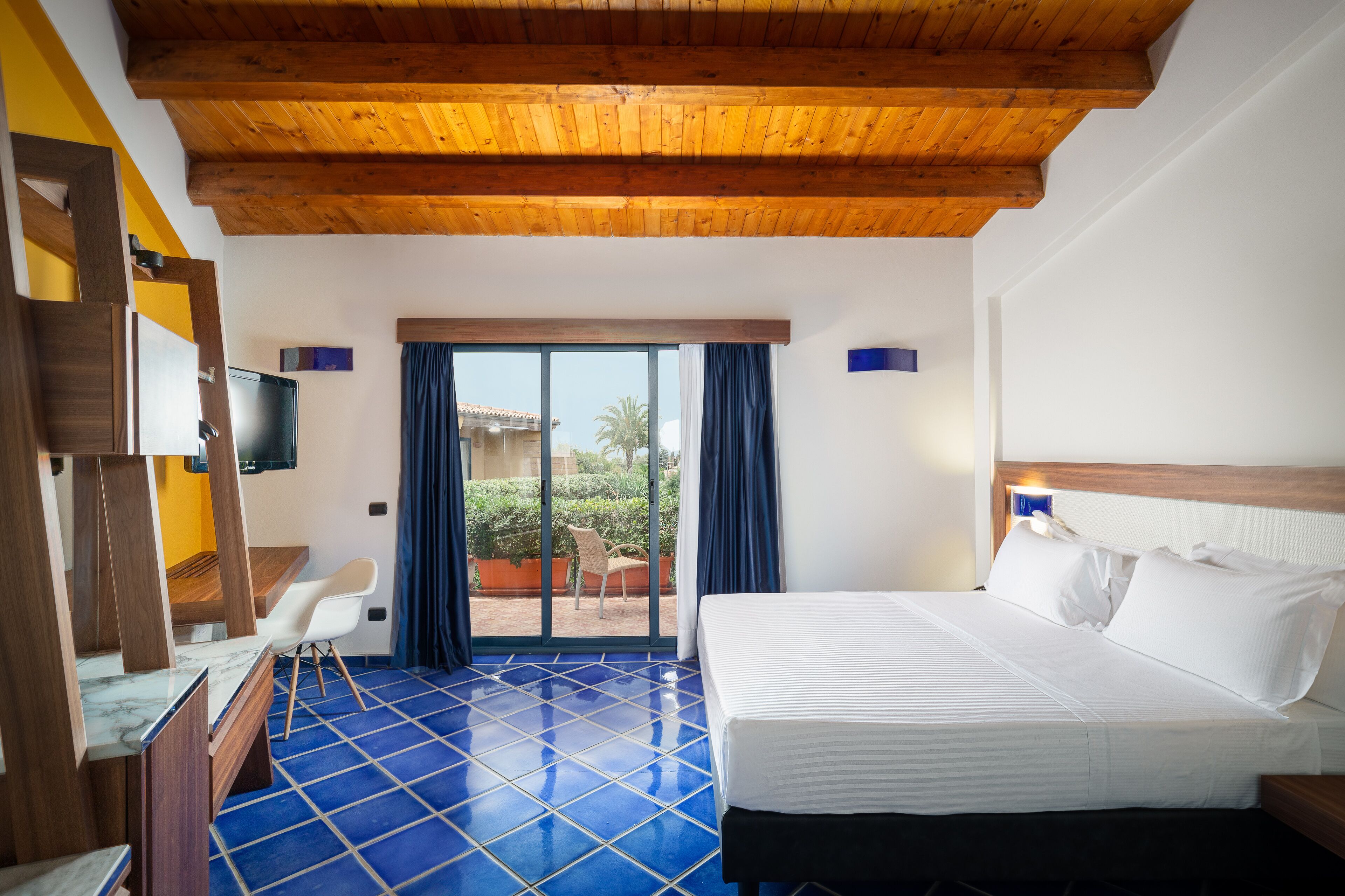 A hotel room with a wooden ceiling, a bed with white linens, a blue-tiled floor, a TV on the wall, and a view of the outdoors through a sliding door.