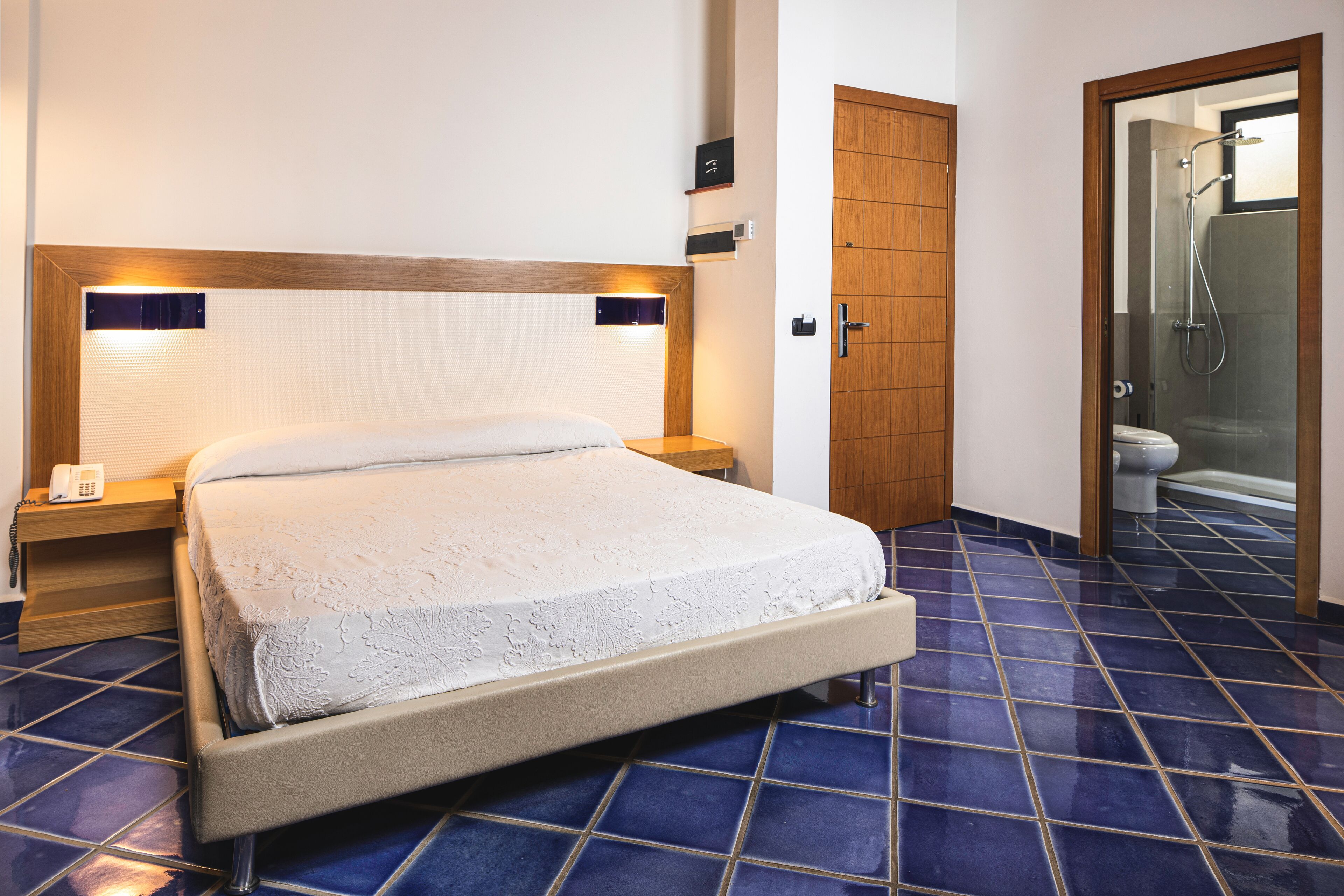 A hotel room with a bed, a bedside table with a phone, a wooden door, and a shower area.