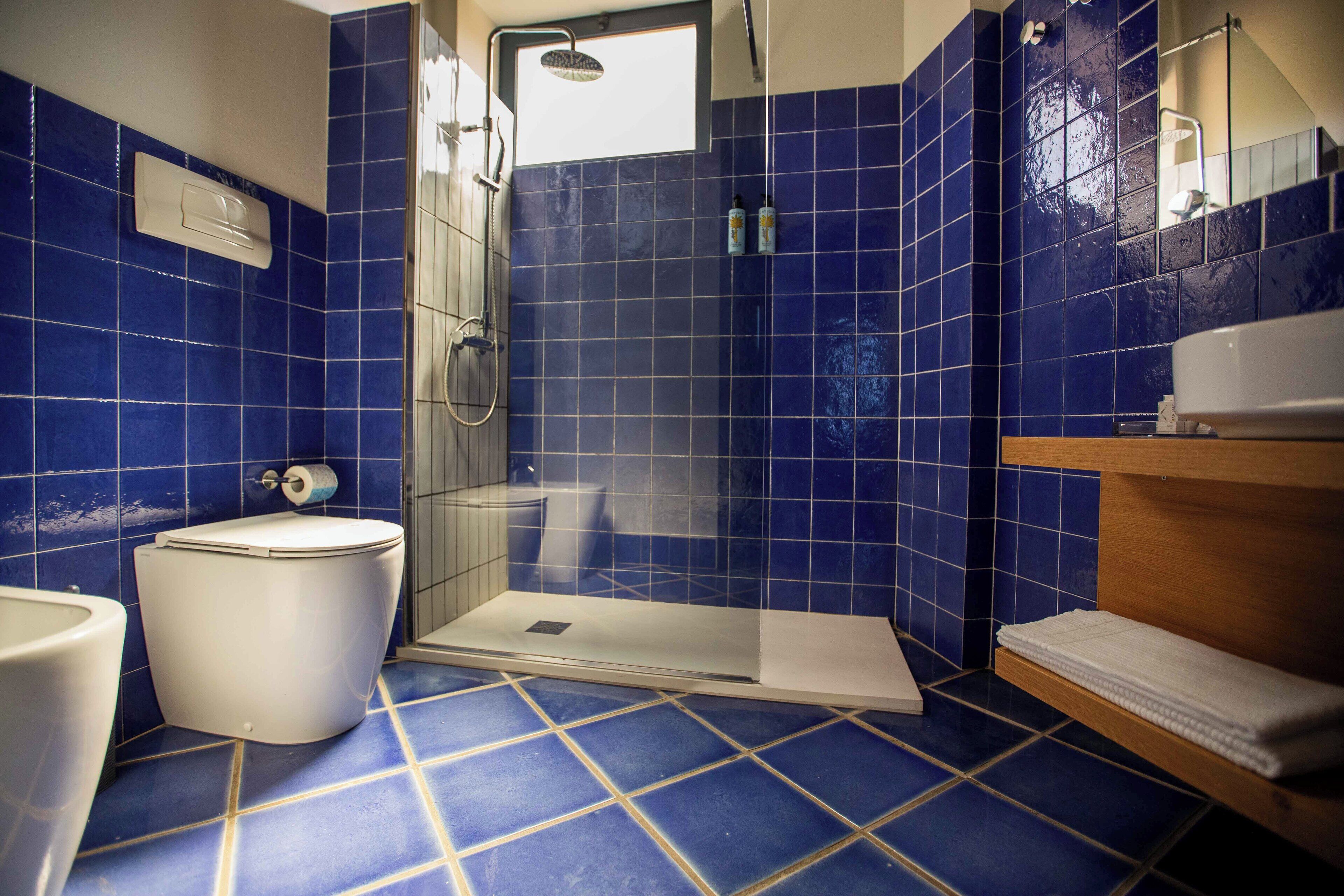 Une salle de bain avec des carreaux bleus, des toilettes blanches, un espace douche et un meuble-lavabo en bois doté d’un porte-serviettes.