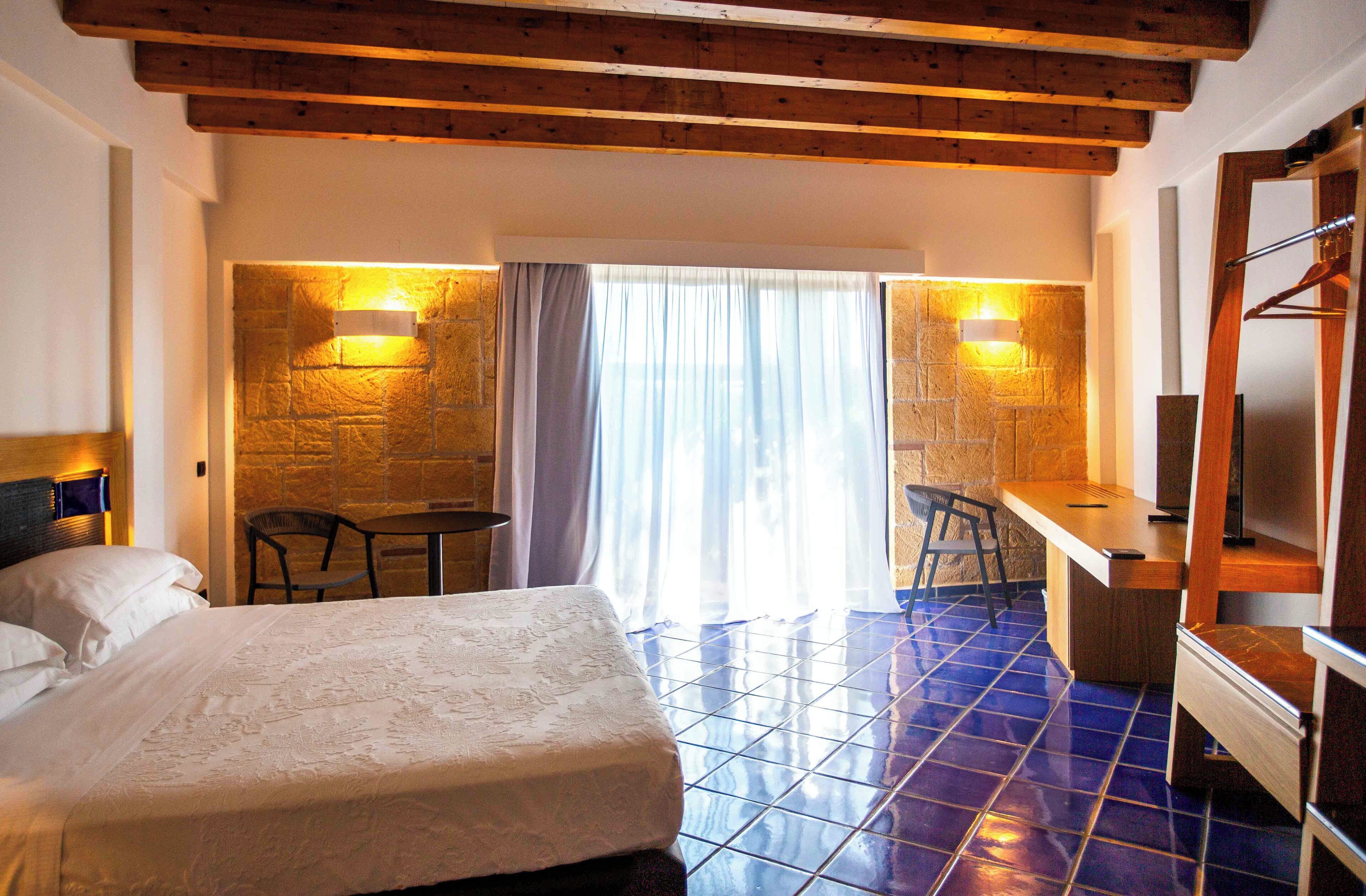 A hotel room with a bed, a desk, and a chair. There are two wall-mounted lights and a window with curtains.