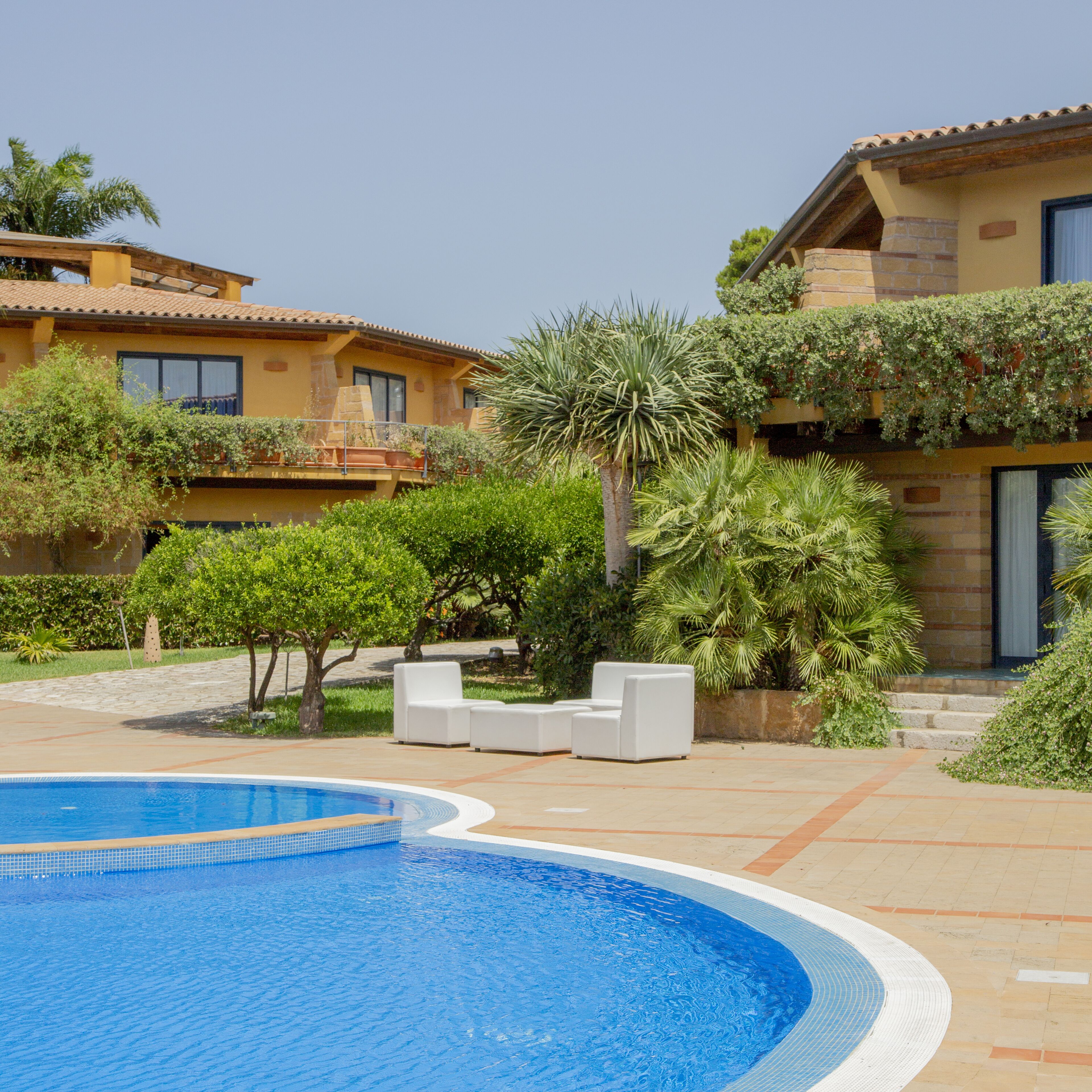 A pool area with a clear blue pool, white lounge chairs, and a building with a tiled roof and greenery.