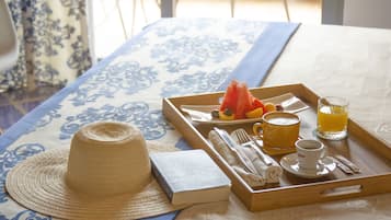 Bandeja de desayuno con sombrero de paja, un libro y una selección de comida que incluye fruta y un vaso de zumo de naranja sobre una cama con edredón estampado.