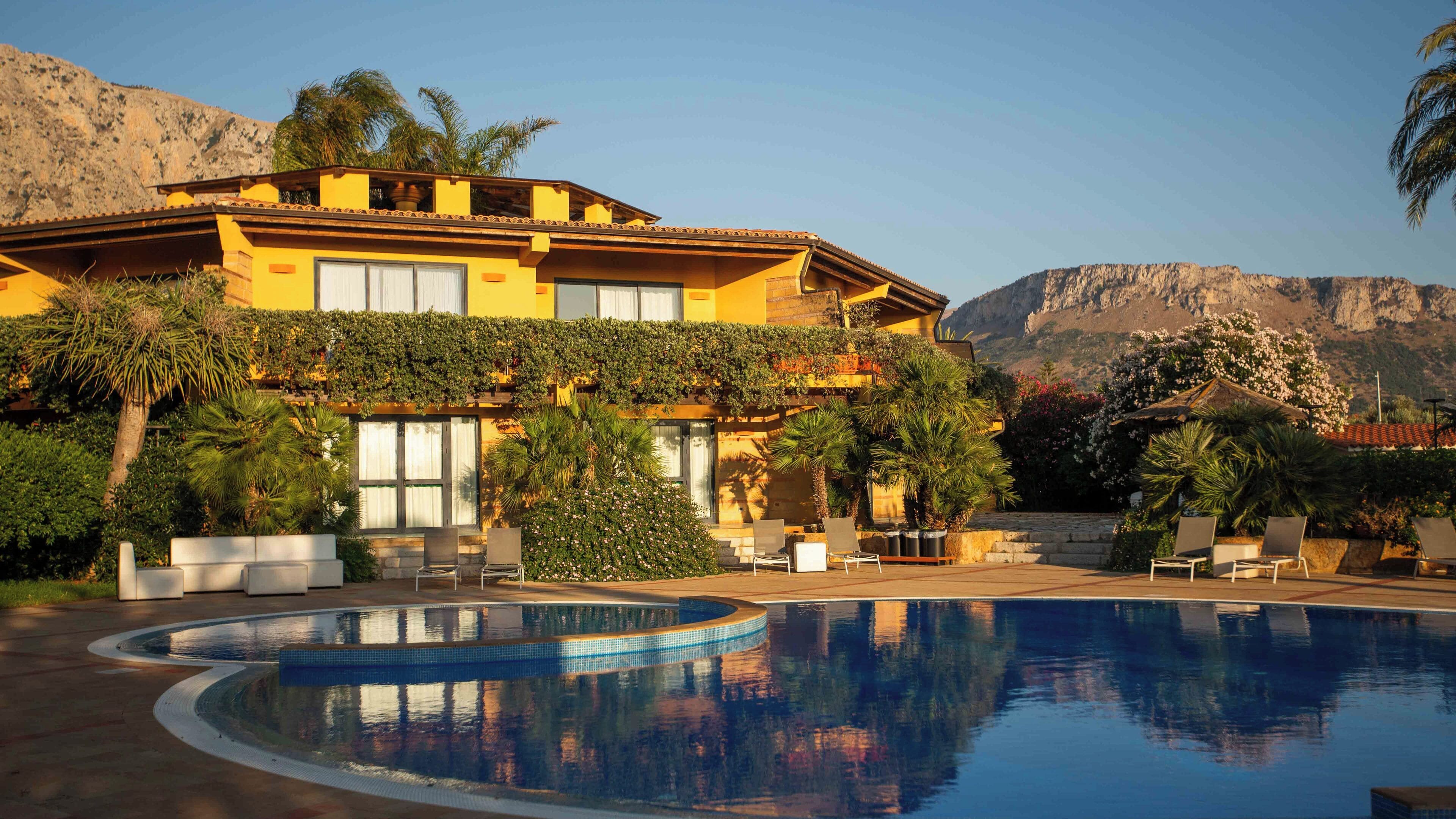 Un edifici gran i groc amb piscina, envoltat de palmeres i un paisatge de muntanyes al fons.