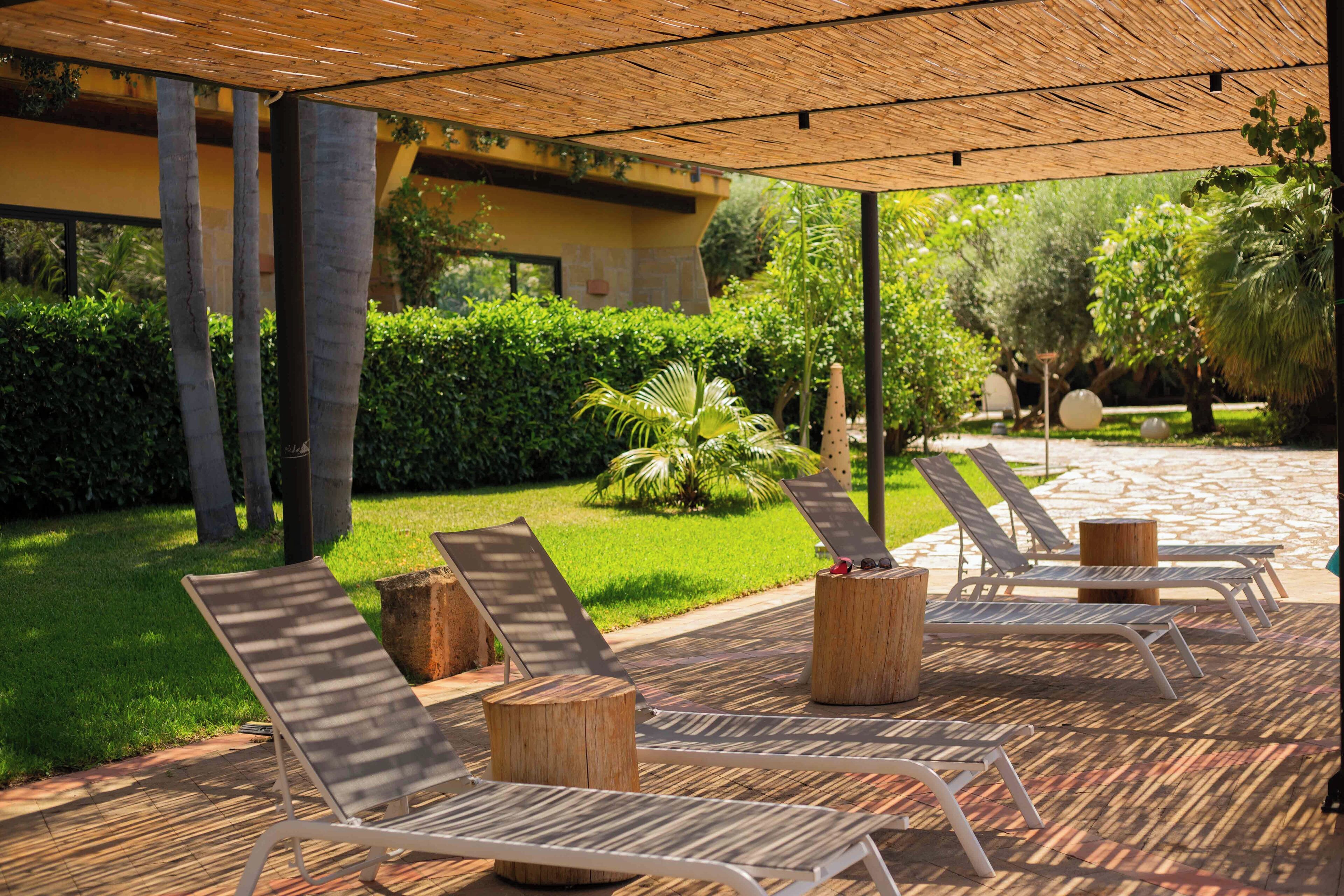A covered outdoor seating area with lounge chairs and wooden stools, surrounded by greenery and a building in the background.