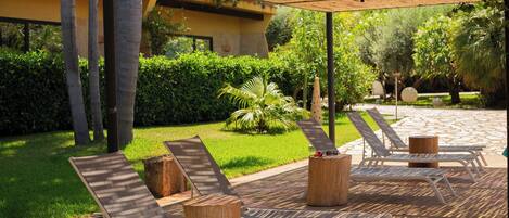 Un espacio al aire libre con techo, con sillones de descanso y taburetes de madera, rodeado de follaje y un edificio al fondo.