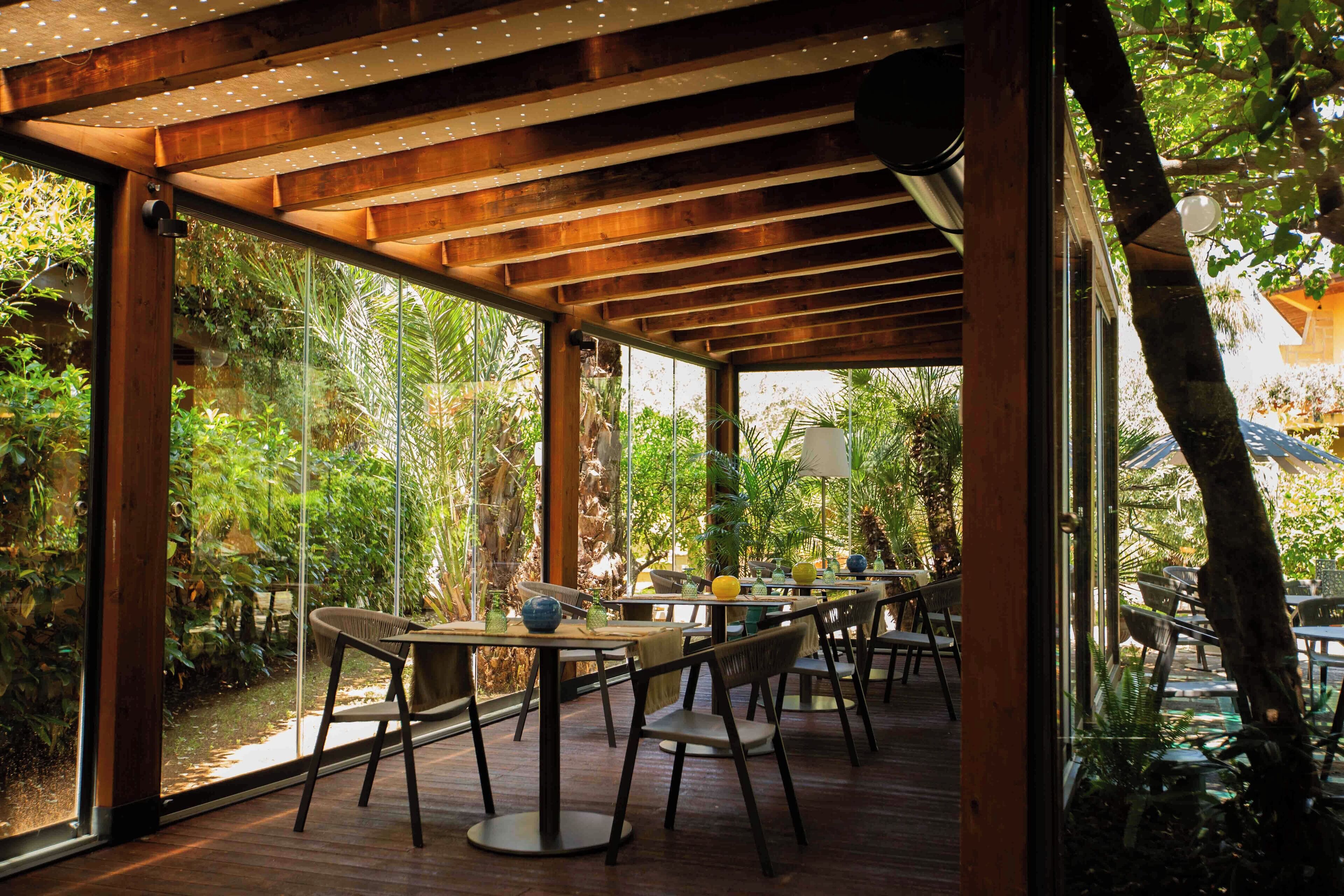 An indoor dining area with wooden tables and chairs, large windows, and a view of greenery outside.