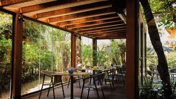 An indoor dining area with wooden tables and chairs, large windows, and a view of greenery outside.