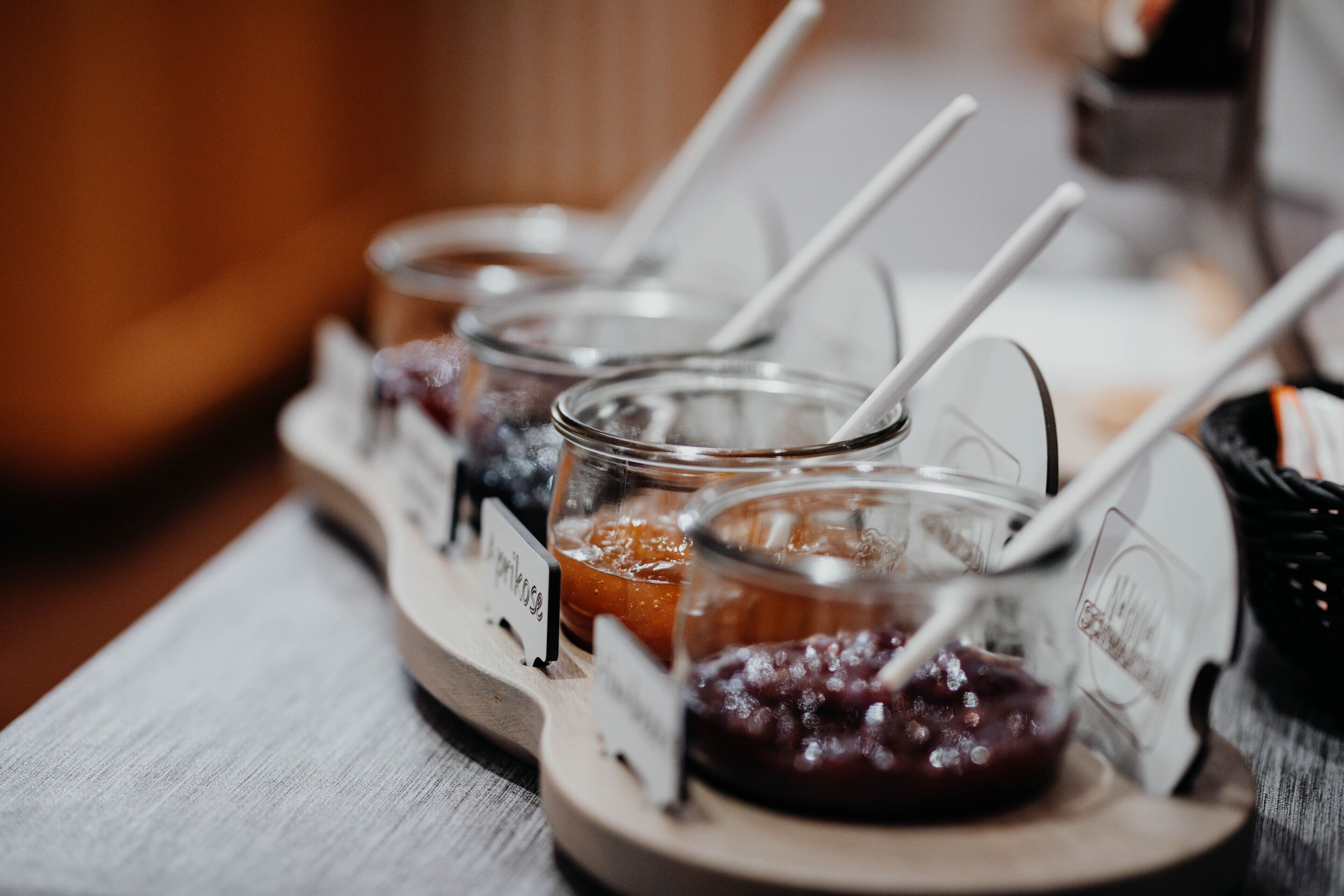 A wooden serving tray with glass containers labeled with names, each containing a different type of food and a spoon.