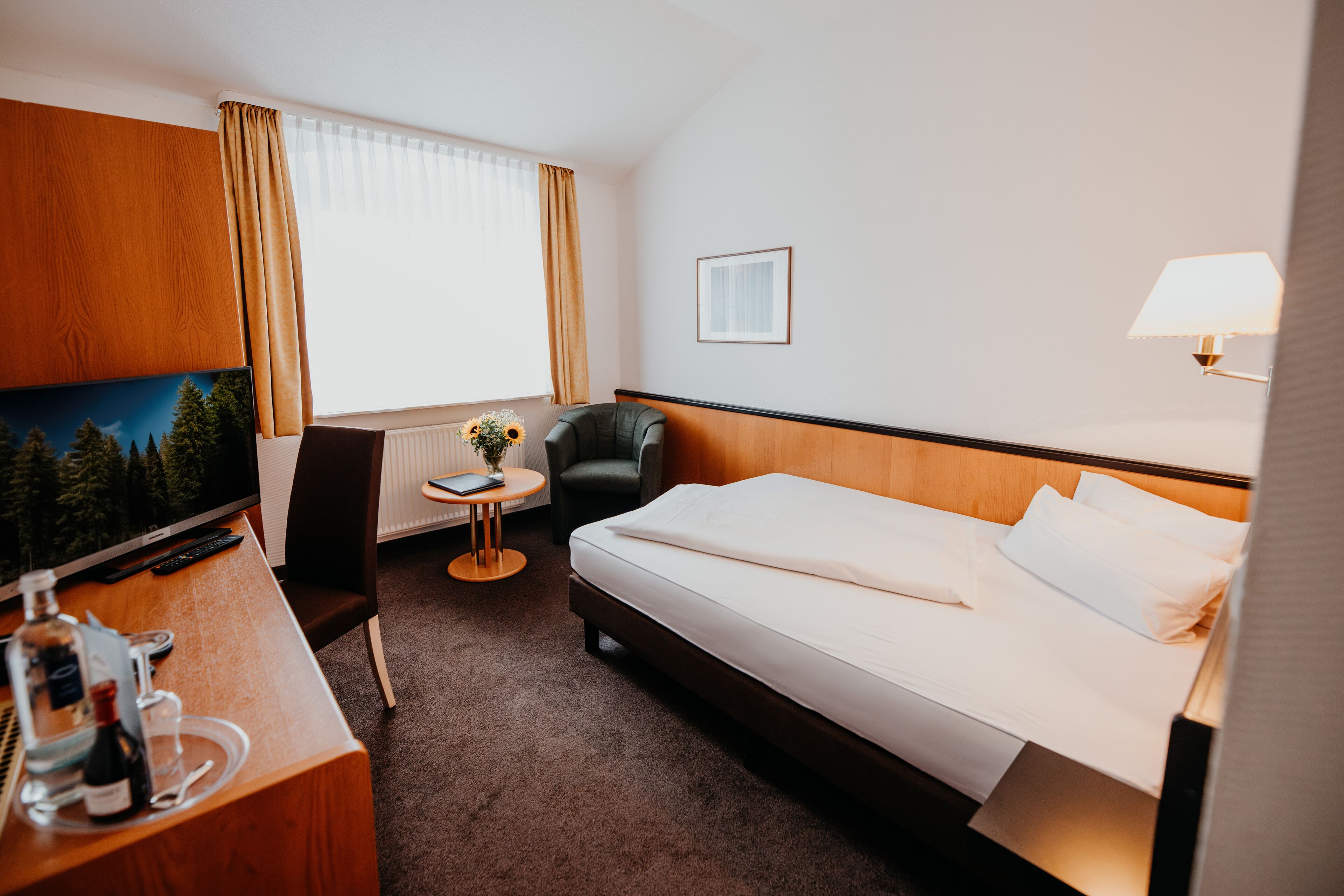 Ein Hotelzimmer mit einem Bett, einem Schreibtisch mit Fernseher, einem Sessel, einem kleinen Tisch mit Blumenvase und einem Fenster mit Vorhängen.