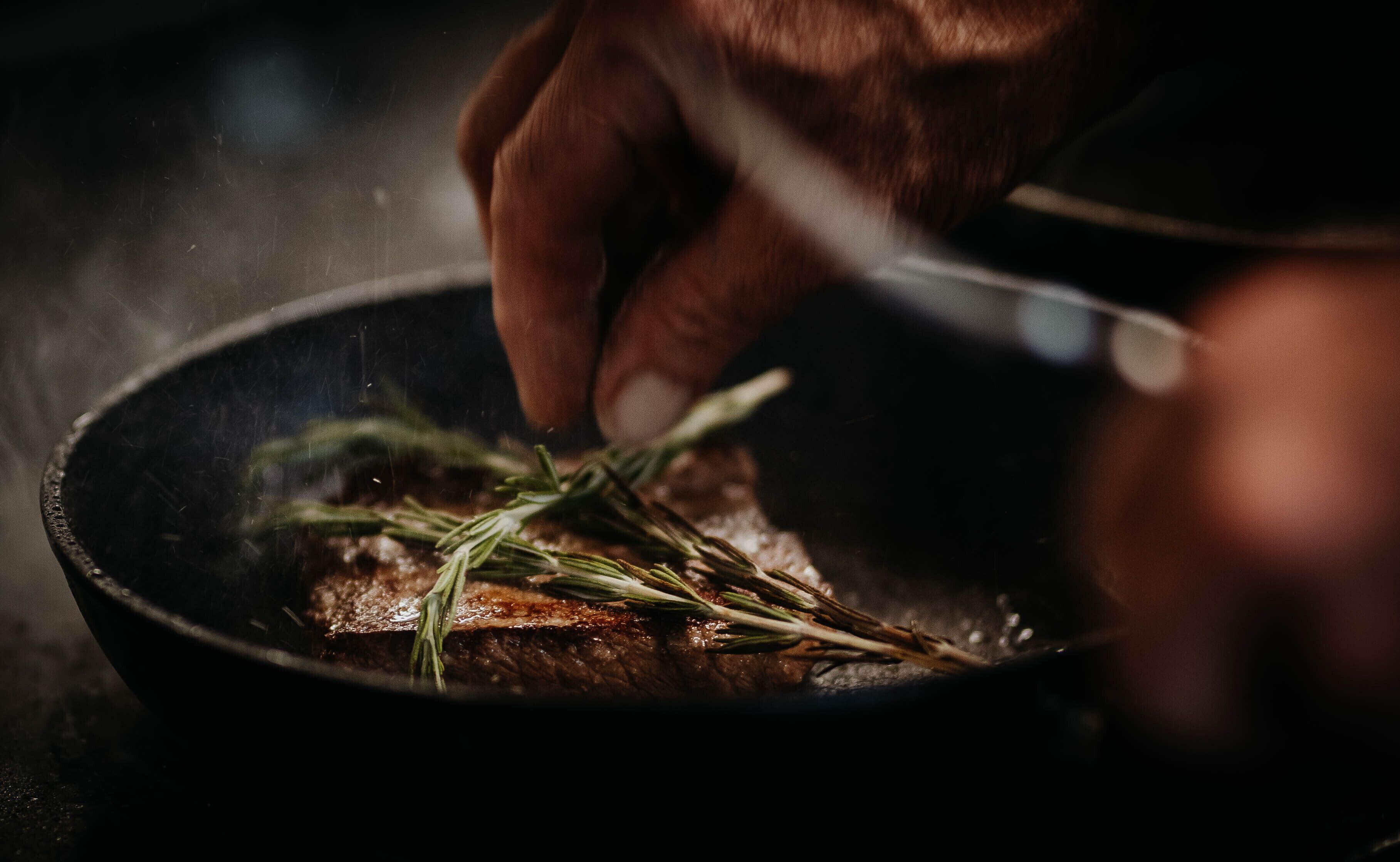 A hand holding a knife over a pan with a steak and rosemary.