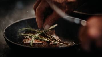 A hand holding a knife over a pan with a steak and rosemary.