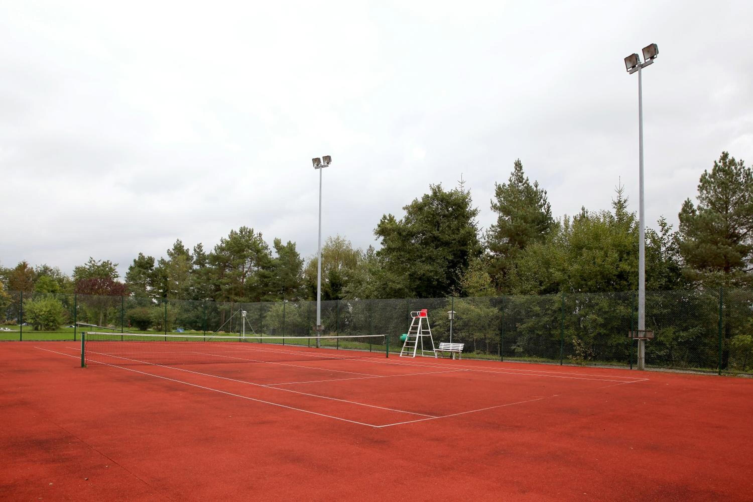 An outdoor tennis court with a net, two floodlights, a ladder, and a bench.