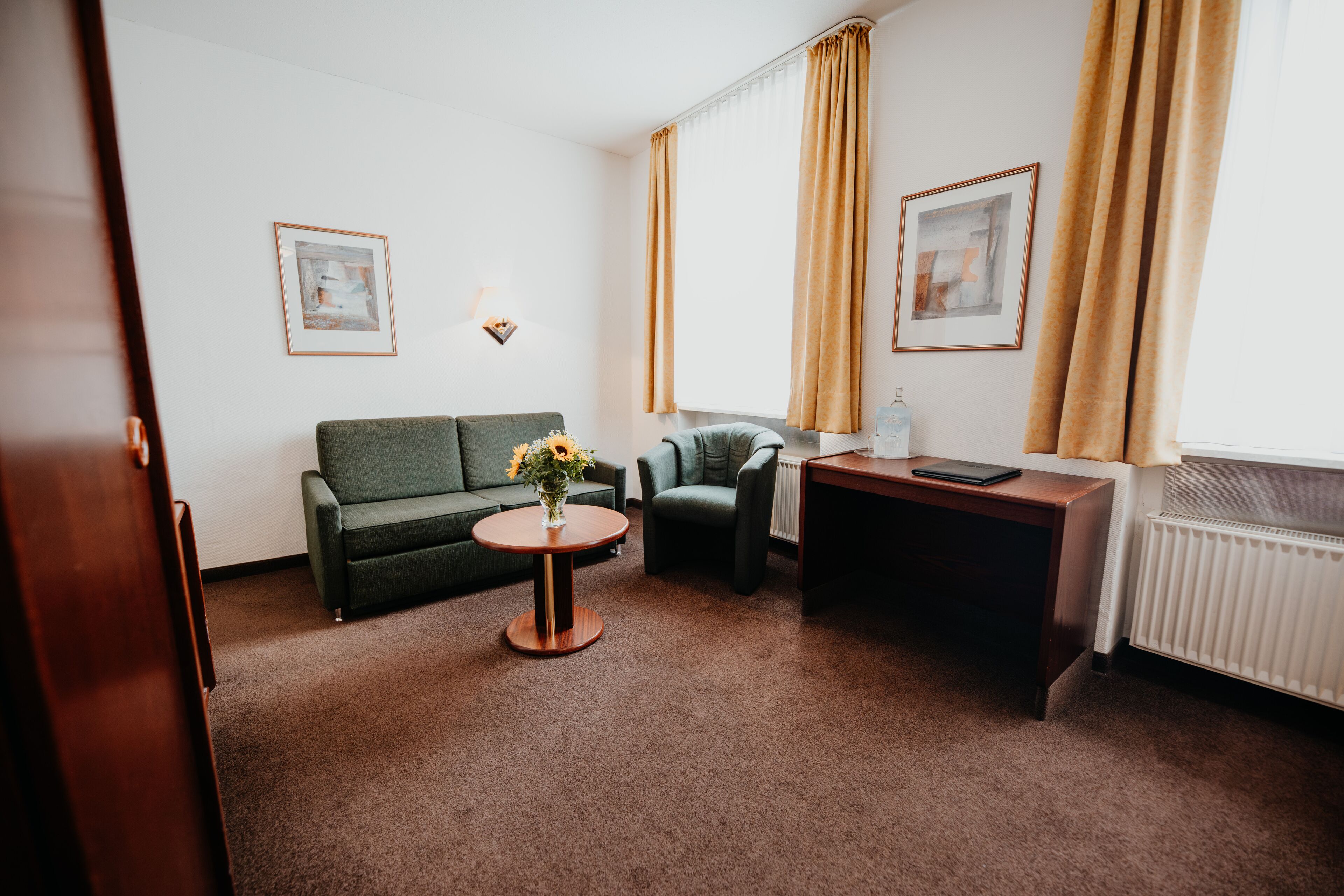 A hotel room with a green sofa, a small round table with a vase of flowers, a desk with a laptop, and two framed pictures on the wall.