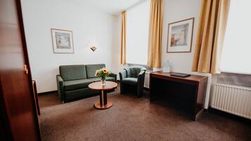 A hotel room with a green sofa, a small round table with a vase of flowers, a desk with a laptop, and two framed pictures on the wall.