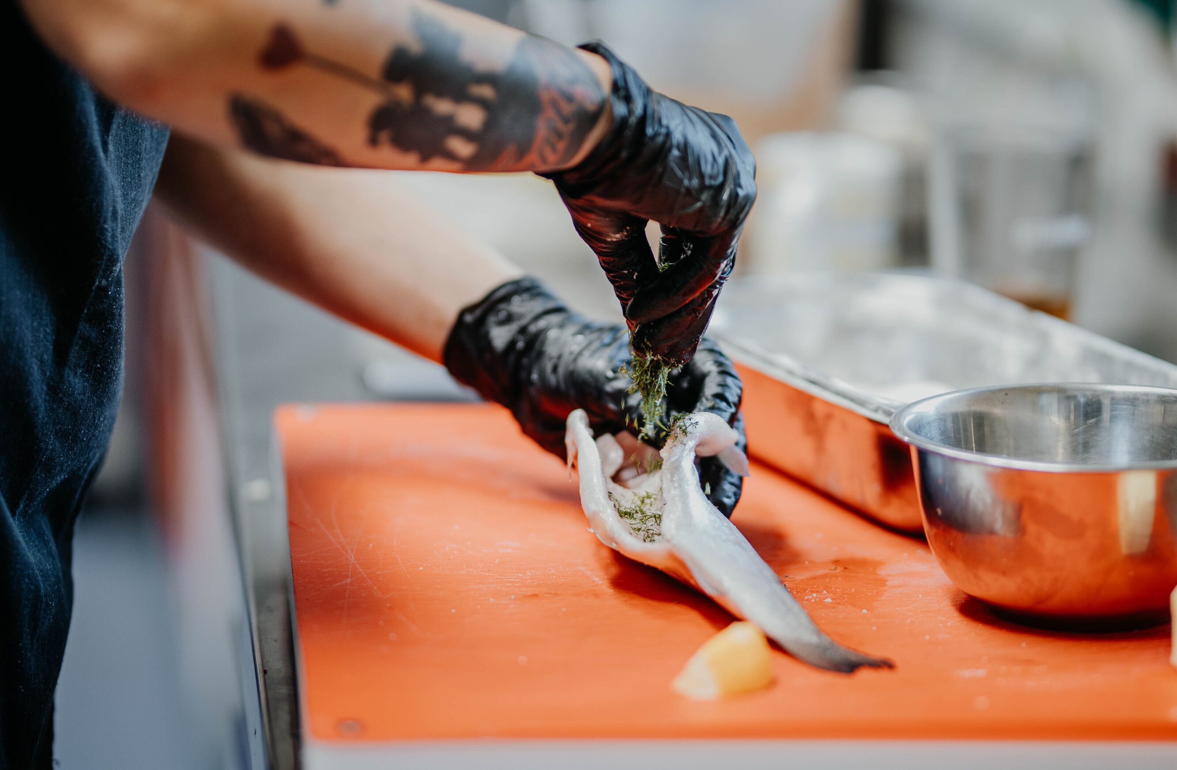 Una persona preparando comida con un cuchillo sobre una tabla de cortar naranja, usando guantes negros, y un recipiente metálico cerca.