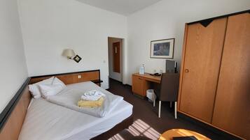 A hotel room with a bed, a desk, a chair, a wardrobe, and a small table.