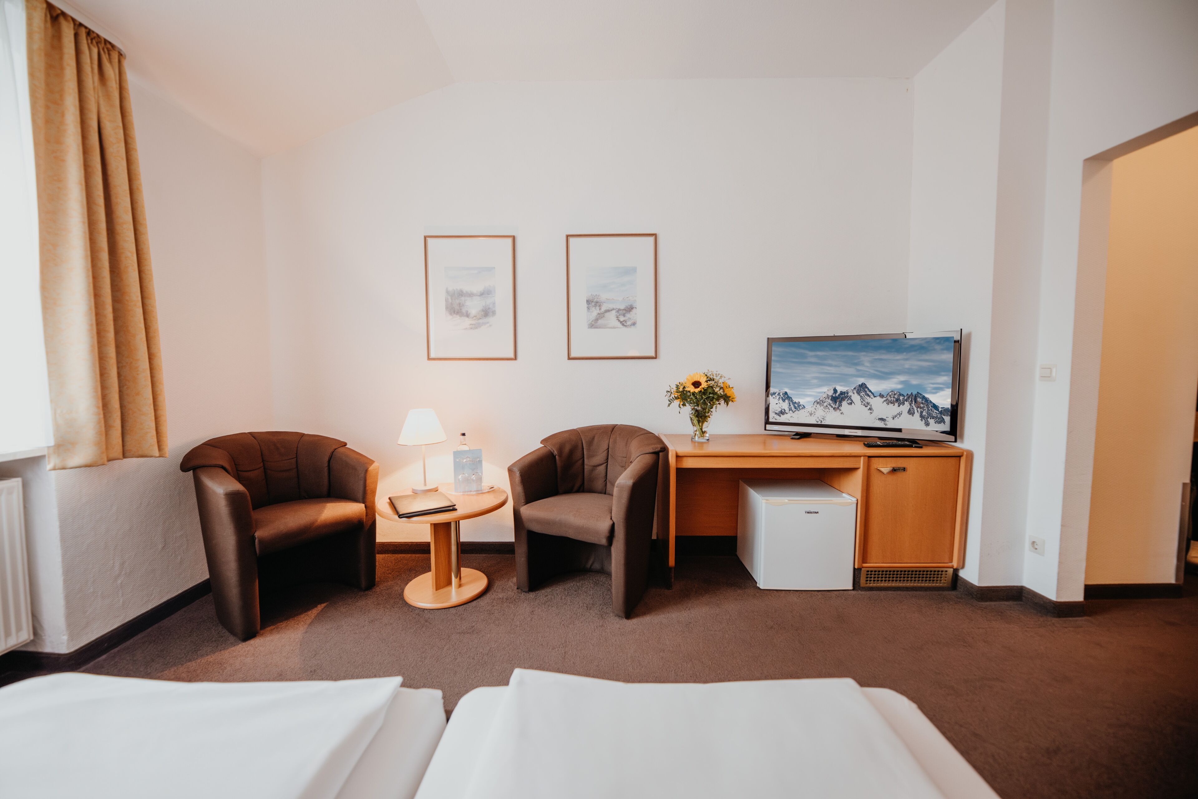 Una habitación de hotel con dos sillones de cuero marrón, un escritorio de madera con televisor, una mesita con lámpara y una cama con sábanas blancas.