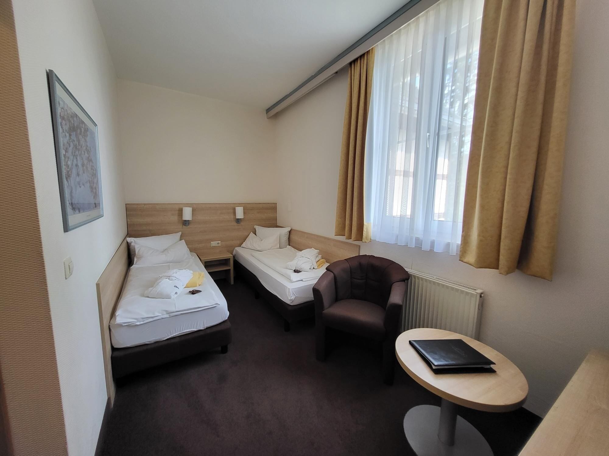 Ein Hotelzimmer mit zwei Einzelbetten, einem kleinen Tisch, einem Stuhl und einem Fenster mit Vorhängen.