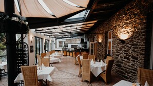 An outdoor dining area with wicker chairs and white tablecloths, a stone wall, and a kitchen in the background. - Sonnenhotel Hoher Hahn (Schwarzenberg/Erzgeb.)