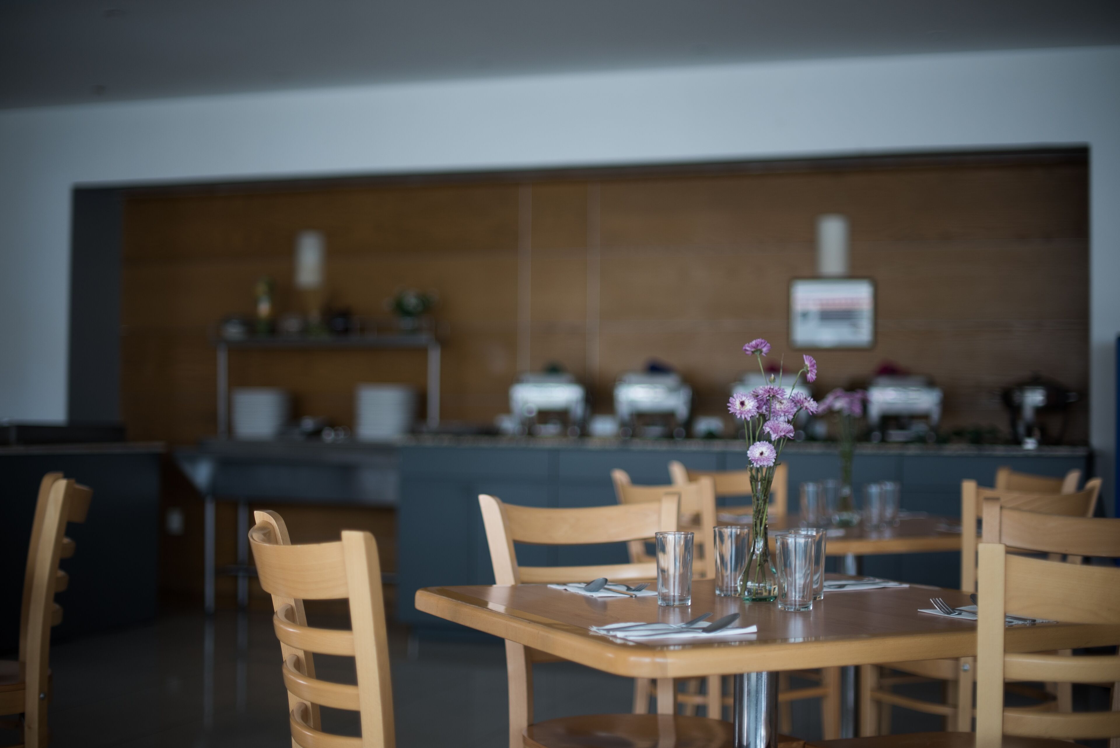 A restaurant with wooden tables and chairs, a vase with flowers, and a counter in the background.