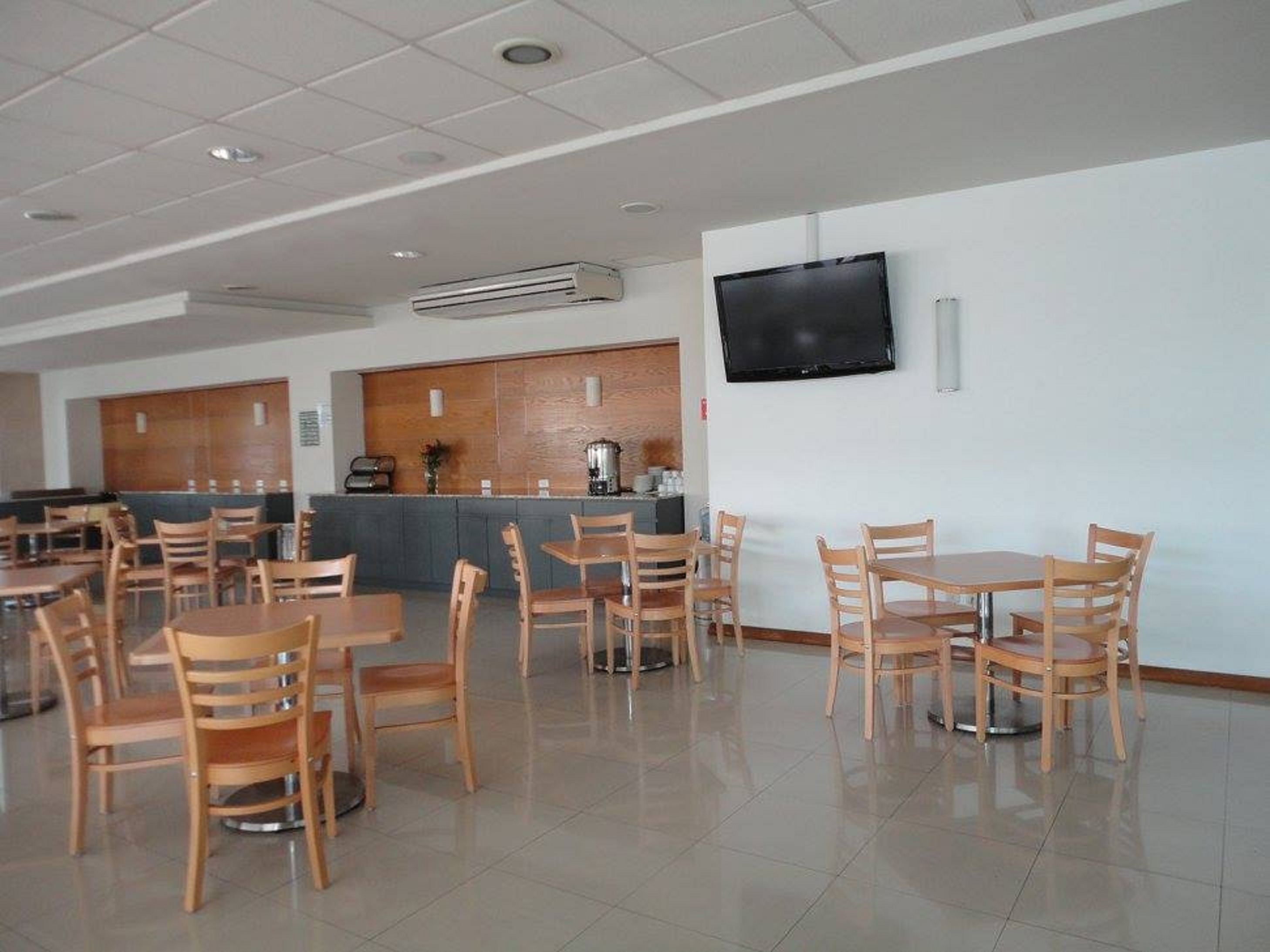 A clean, modern dining area with wooden tables and chairs, a flat-screen TV mounted on the wall, and a coffee machine at the counter.