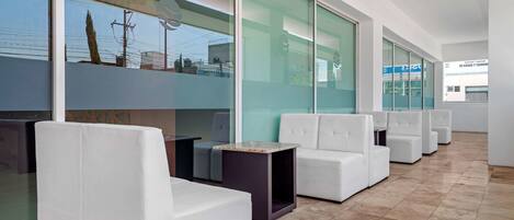 A modern indoor space with white sofas and a tiled floor, large windows, and a view of the outside.