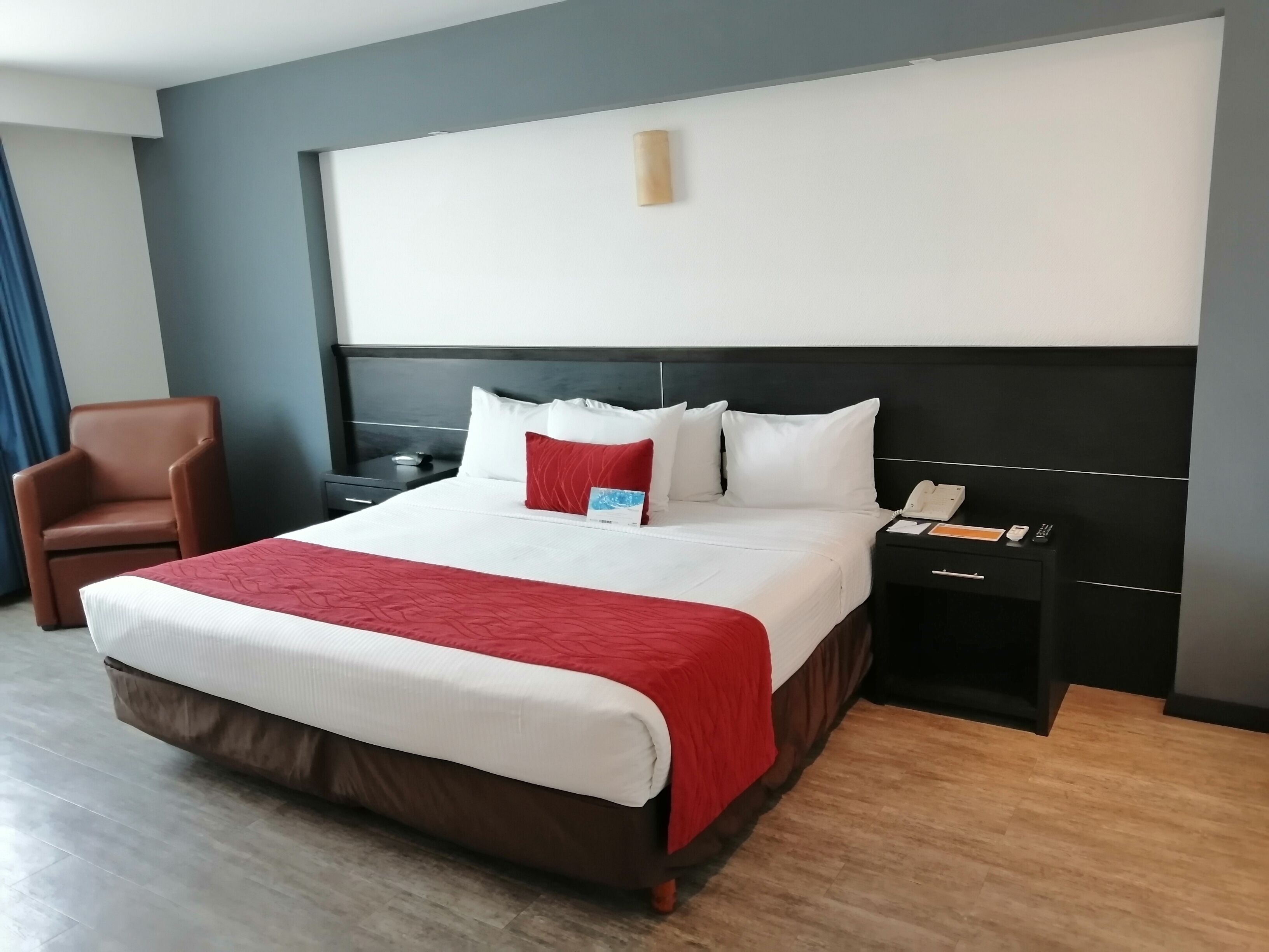 Une chambre d’hôtel avec un grand lit, un fauteuil marron, une table de chevet avec un téléphone et une lampe fixée au mur.