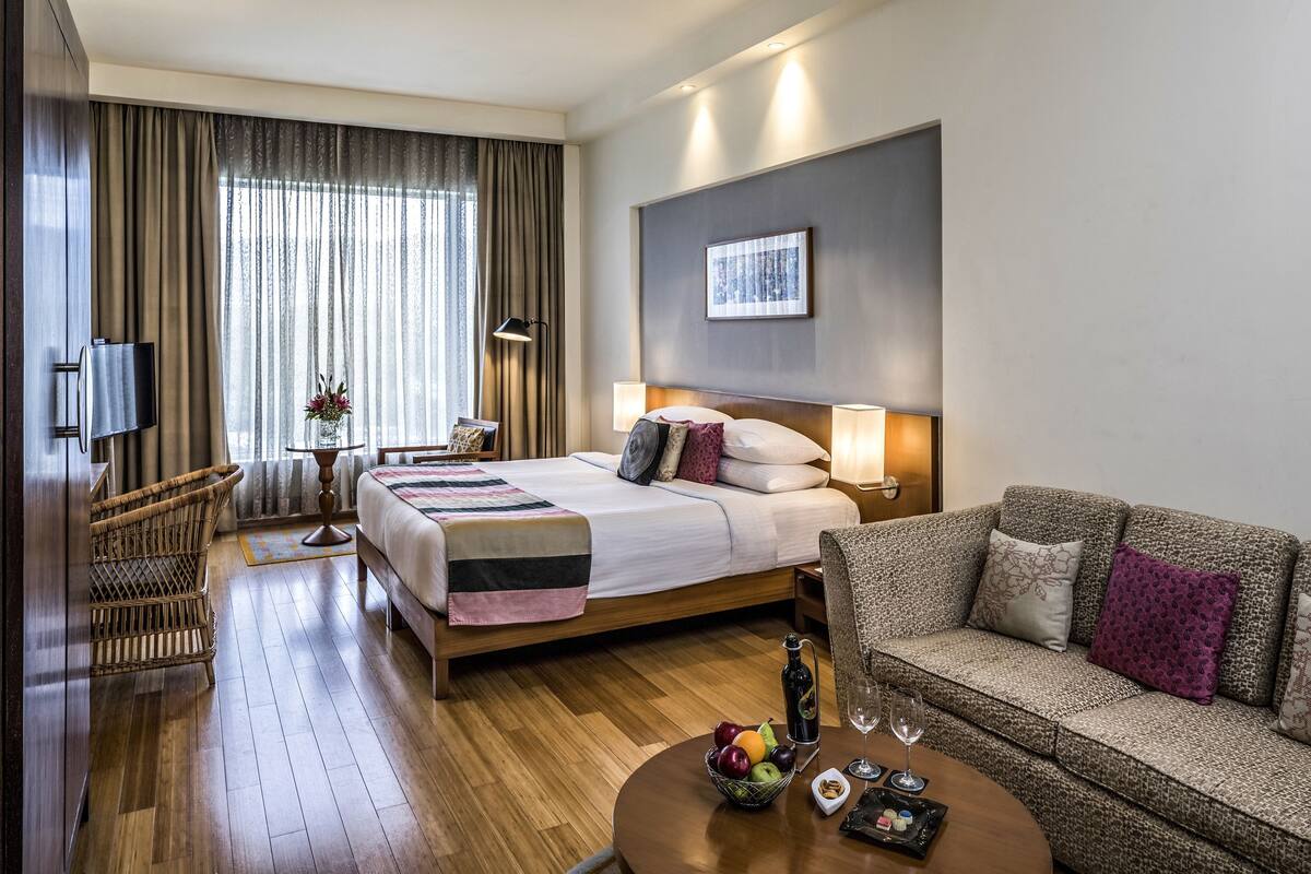A hotel room with a bed, a sofa, a TV, a table with fruit and snacks, and a window with curtains.