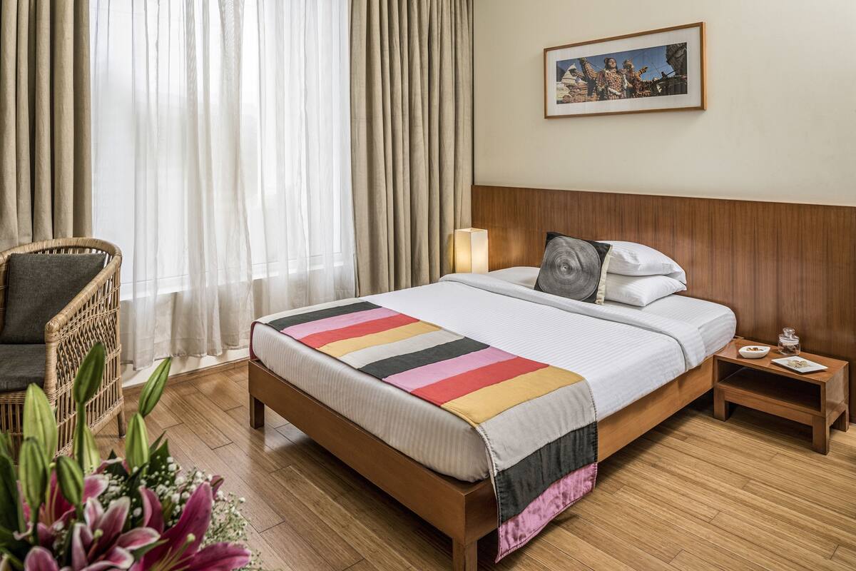 A hotel room with a large bed, a wooden headboard, a wicker chair, a bedside table with a lamp, and a framed picture on the wall.