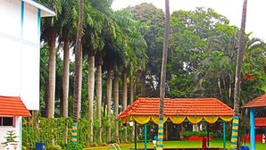 A swimming pool with a clear blue water surface, surrounded by lush greenery and tall palm trees. There are colorful gazebos with red roofs and yellow curtains. - Ramanashree California Resort (Bengaluru)