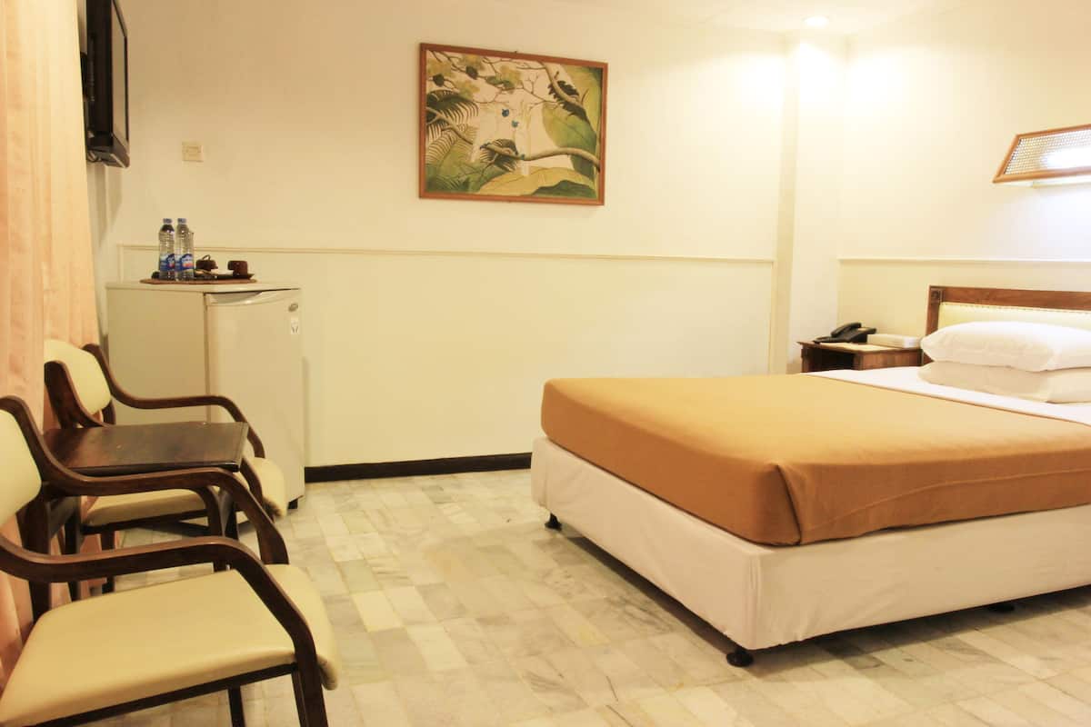 A hotel room with a bed, a desk, a chair, a refrigerator, and a painting on the wall.