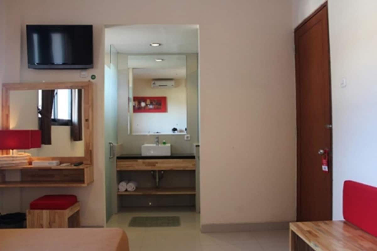 A room with a bed, a desk, a television mounted on the wall, a bathroom with a sink and mirror, and a wooden door.