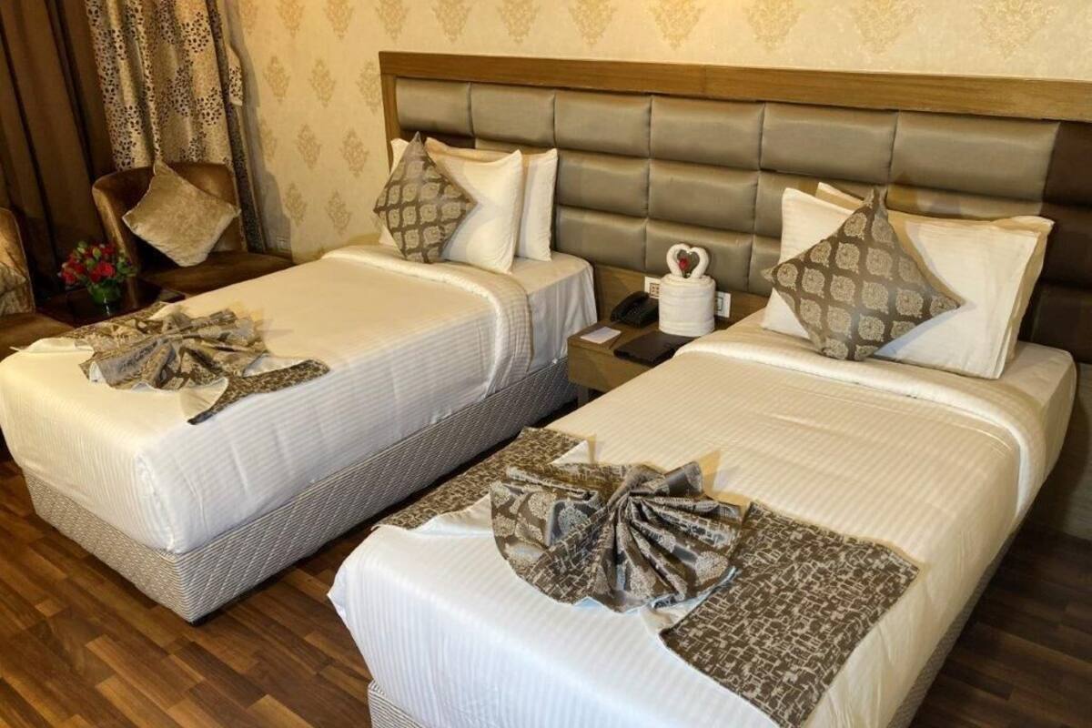 A hotel room with two beds, each with a patterned bedspread and a decorative bow. There is a wooden headboard, a bedside table with a lamp, and a telephone.