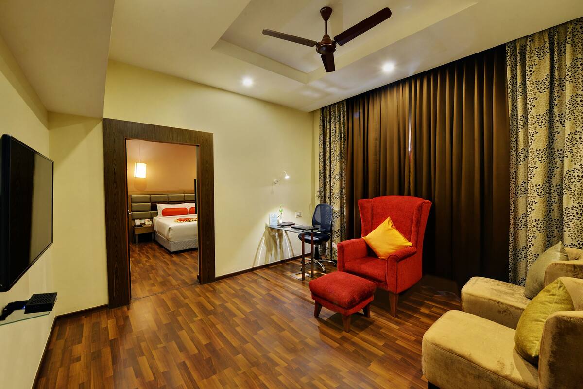 A hotel room with a wooden floor, a red armchair, a beige sofa, a television, and a ceiling fan.