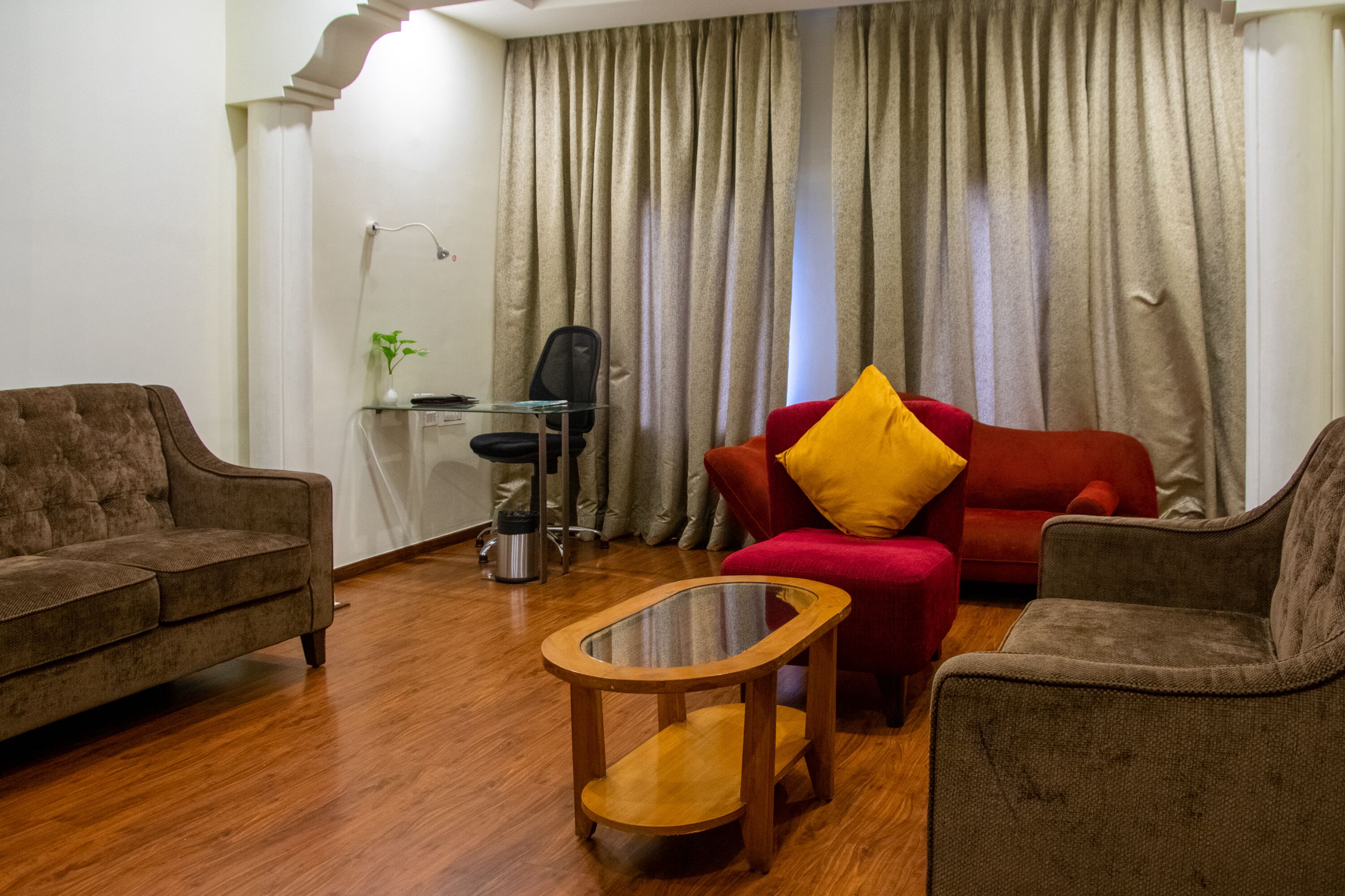 Un salon avec un canapé marron, un fauteuil rouge avec un coussin jaune, une table basse en verre et un bureau avec une chaise et une lampe.