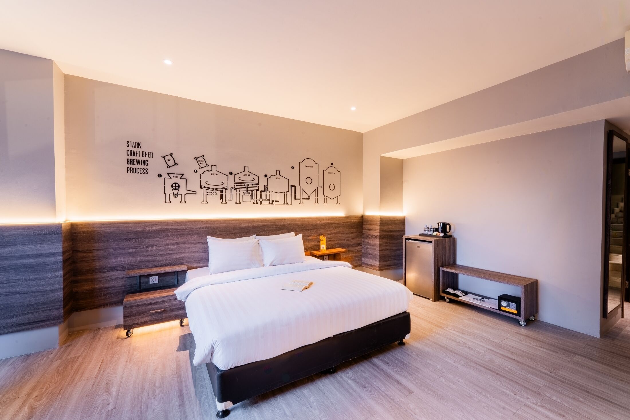 Une chambre d’hôtel moderne dotée d’un grand lit, de tables de chevet, d’une petite table, d’un bureau et d’une œuvre d’art murale représentant le processus de fabrication de la bière artisanale de la marque Stork.