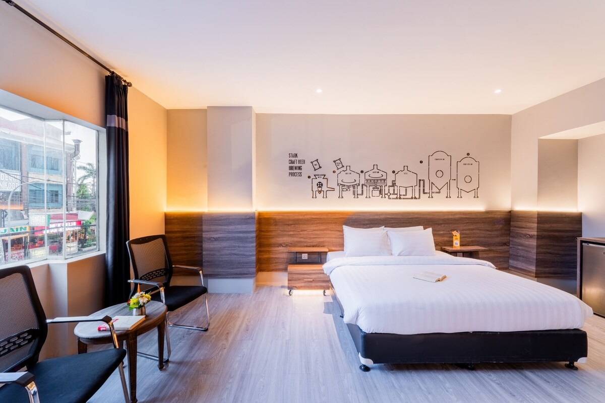 A modern hotel room with a large bed, a desk, and a chair. There is a window with a view of buildings outside.