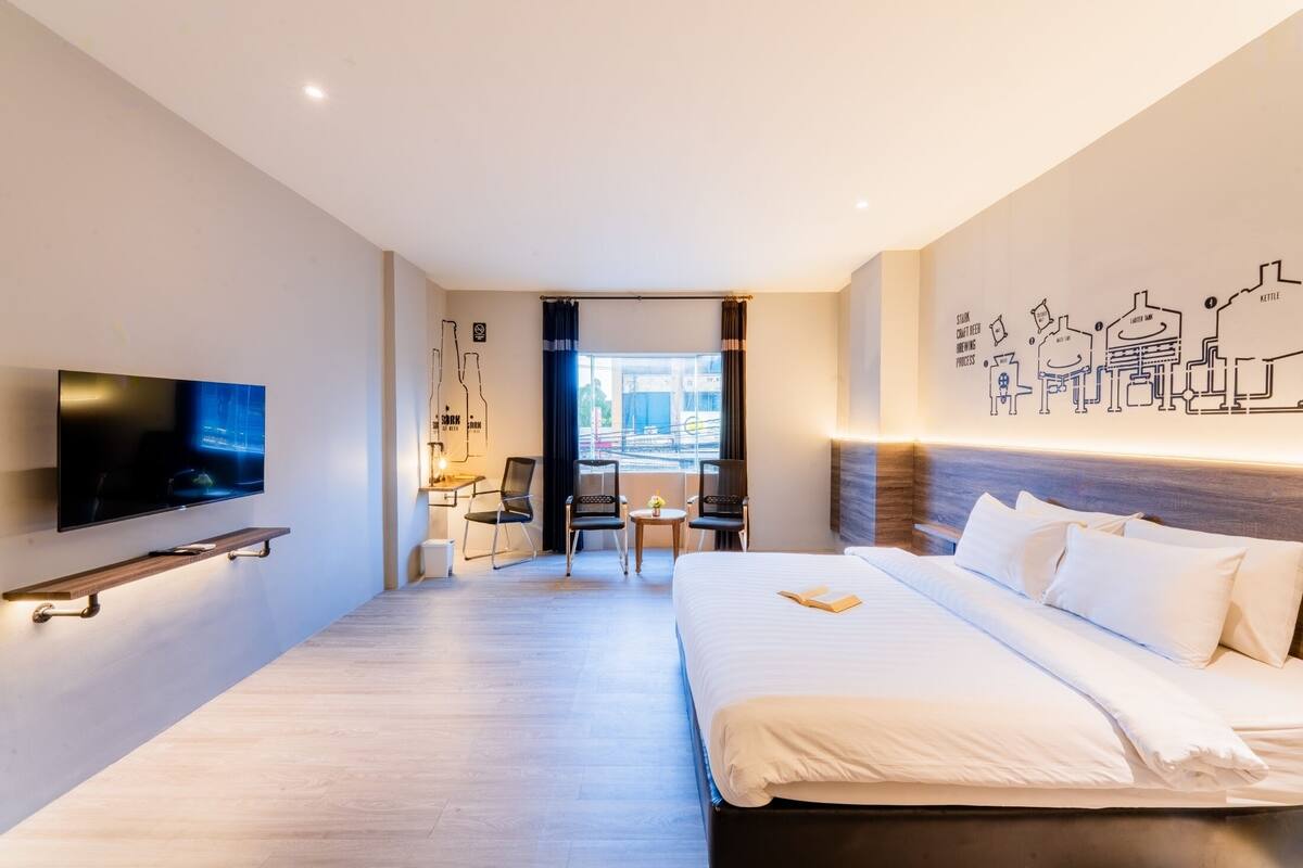 A hotel room with a large bed, a flat-screen TV mounted on the wall, a small desk with a chair, and a view of the city through a window.