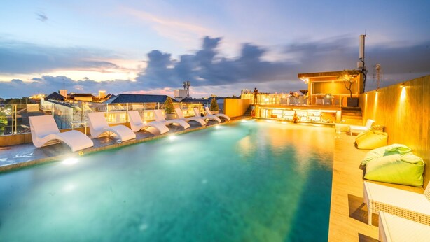 Une piscine dotée de chaises longues et d’un espace bar, le tout entouré de bâtiments au style architectural moderne.