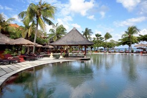 Un espace piscine aménagé dans un complexe hôtelier, avec des chaises longues, un kiosque et des palmiers.
