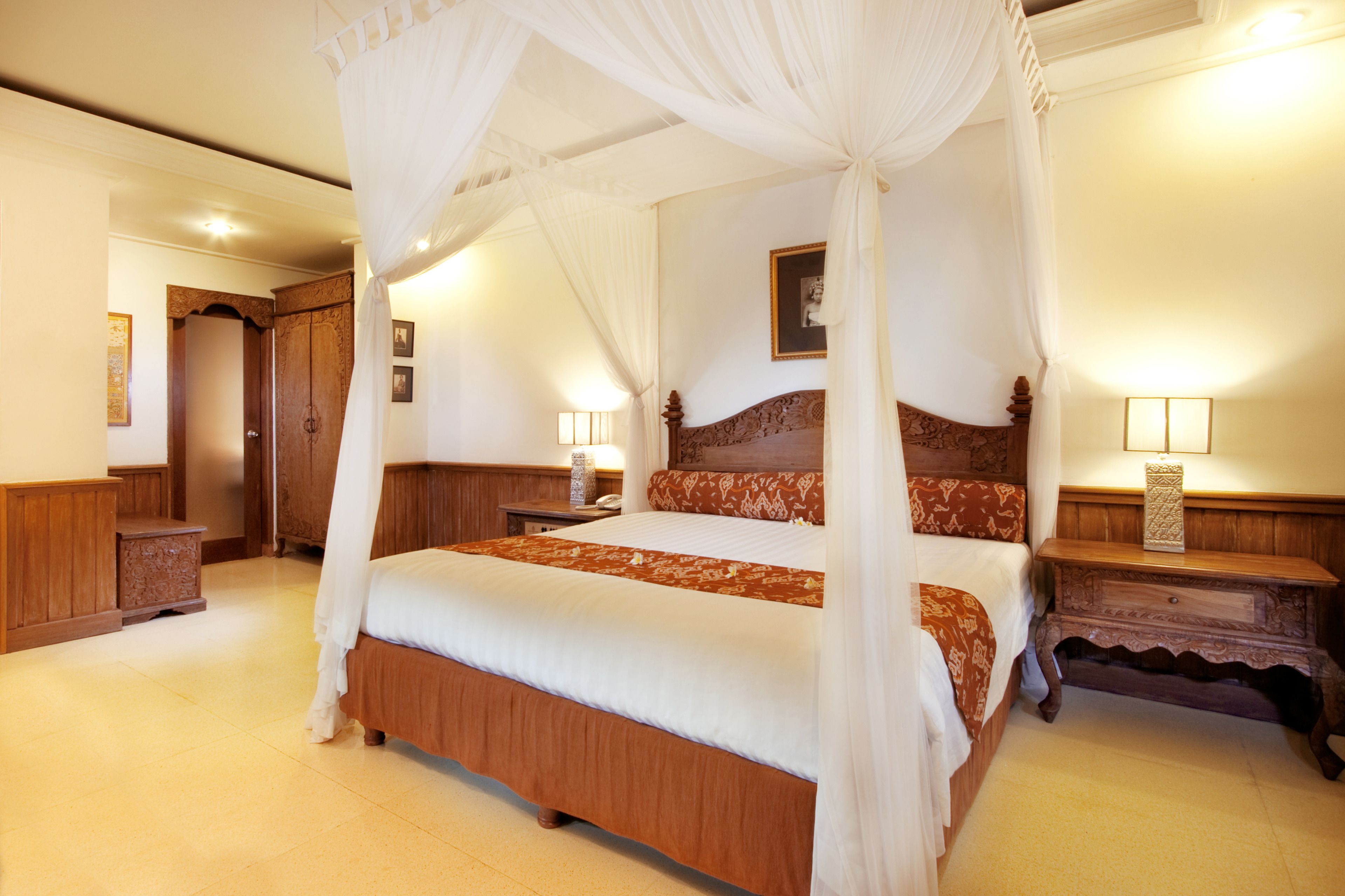 Una cama con dosel sobre cuatro columnas, con cabecera de madera, mesitas de noche con lámparas y un cuadro enmarcado en la pared.