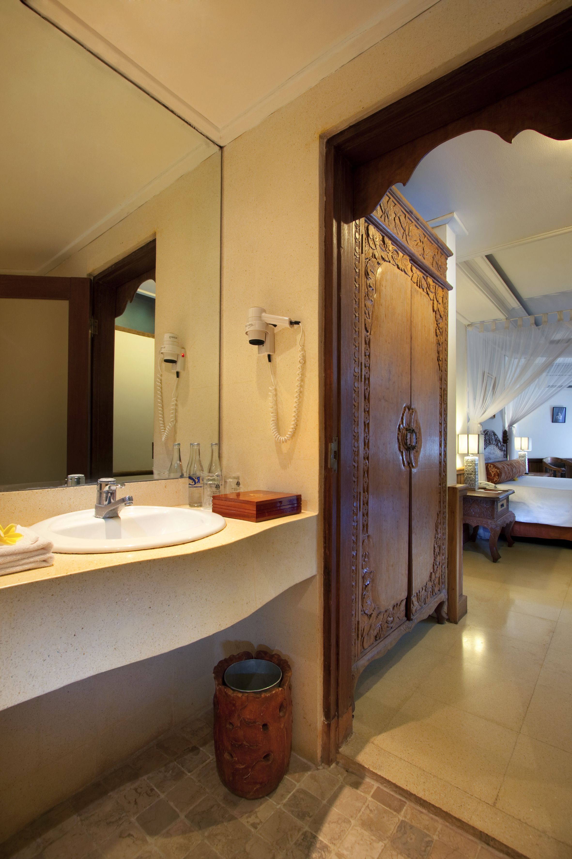 A bathroom with a sink, mirror, and vanity. There is a wooden door with intricate carvings and a small trash can.