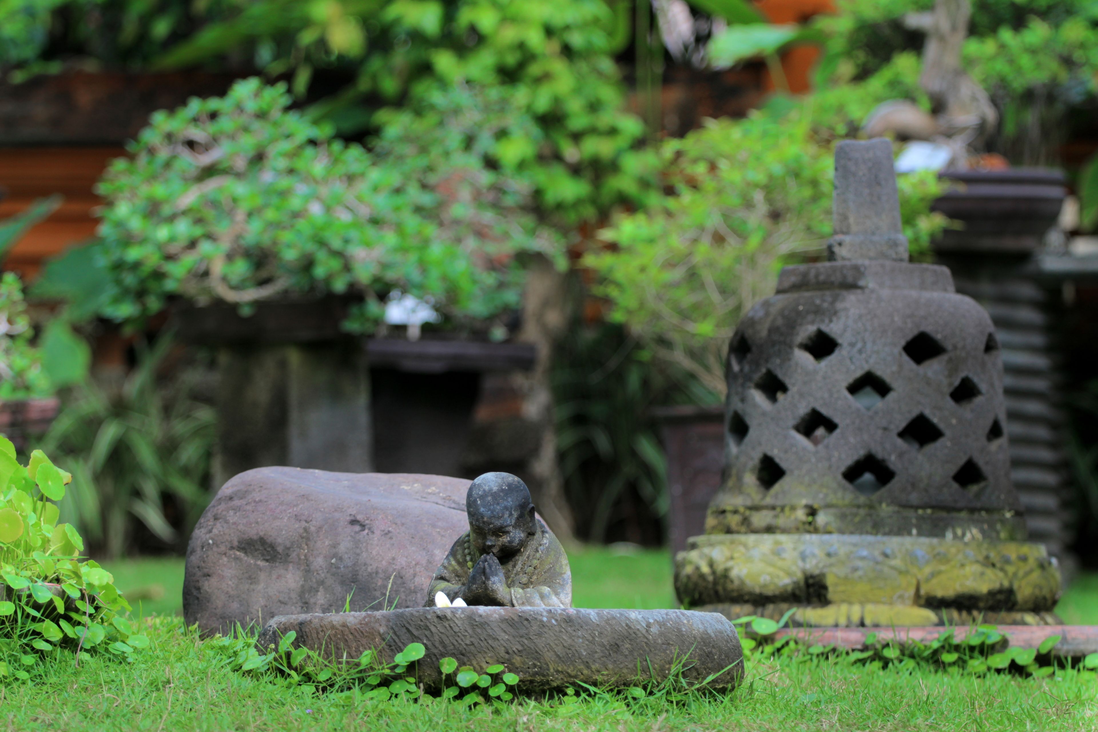 Une statue de pierre représentant une figure, située dans un jardin luxuriant, avec des constructions traditionnelles en arrière-plan.