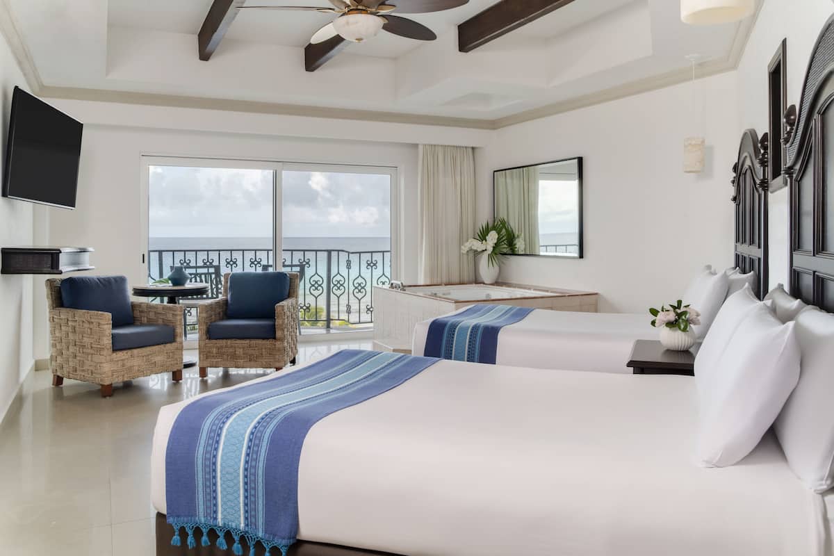 A hotel room with a large bed, two armchairs, a small table, a TV, a balcony with a view, and a ceiling fan.
