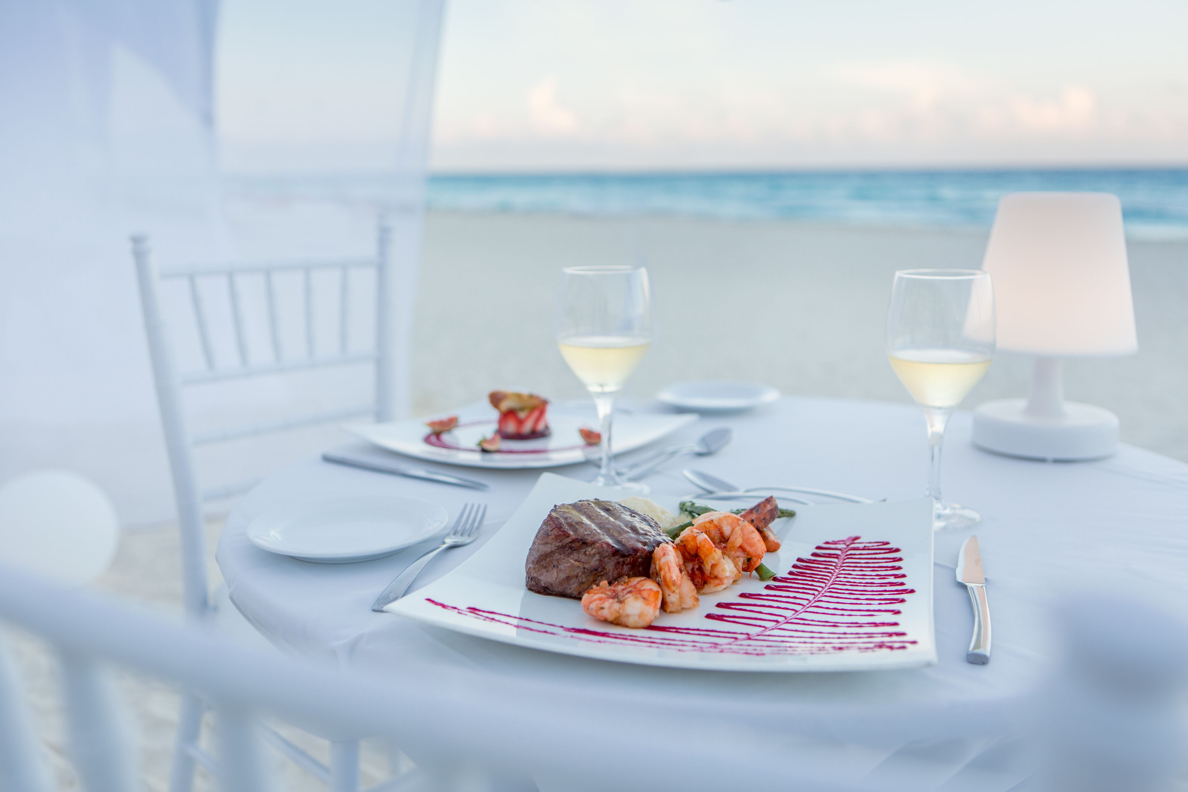 Une table dressée pour deux personnes, avec des fruits de mer et un steak, des verres de vin blanc et une lampe sur une table donnant sur la plage.
