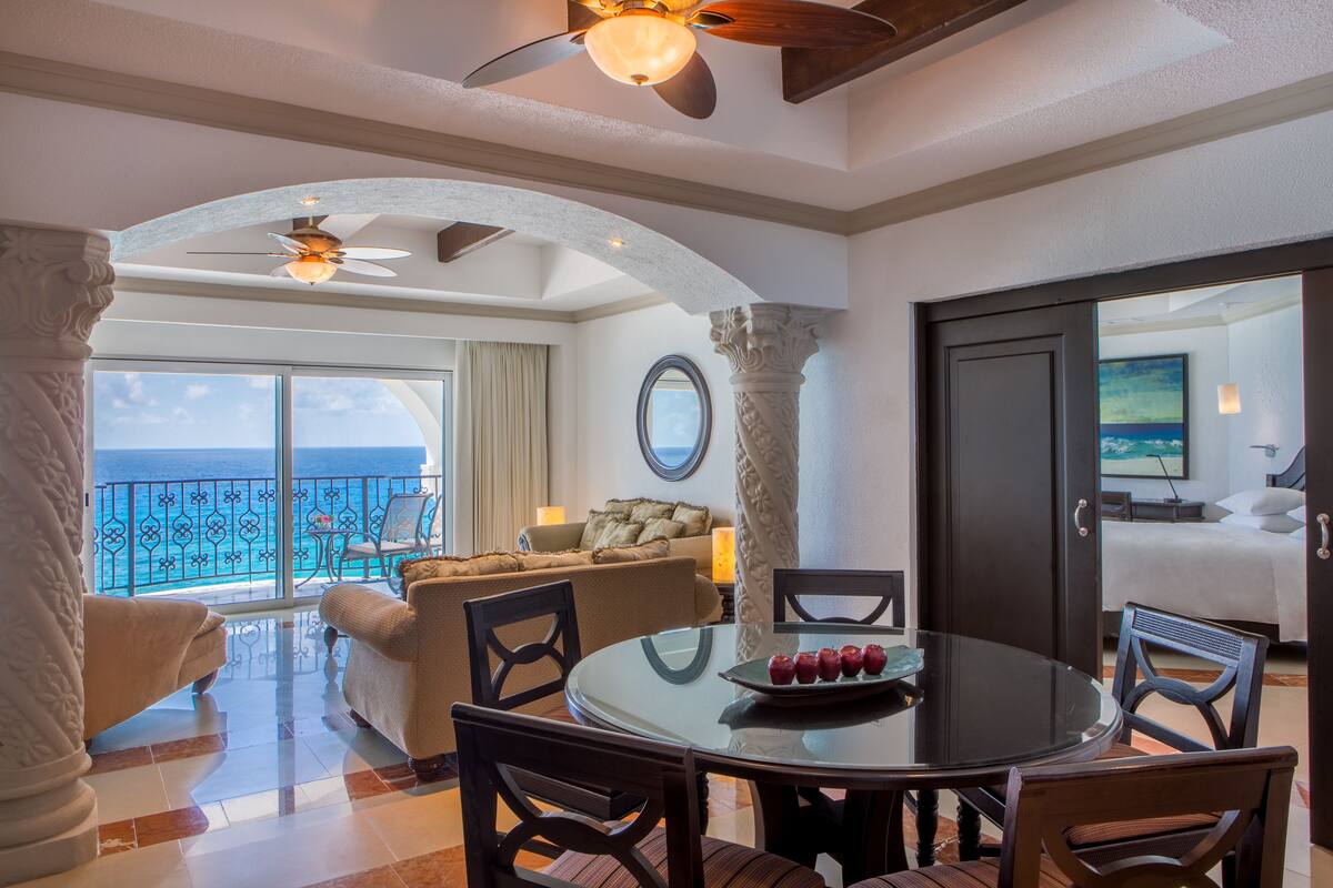 A dining area with a round table, chairs, and a ceiling fan. There is a balcony with a view of the ocean.